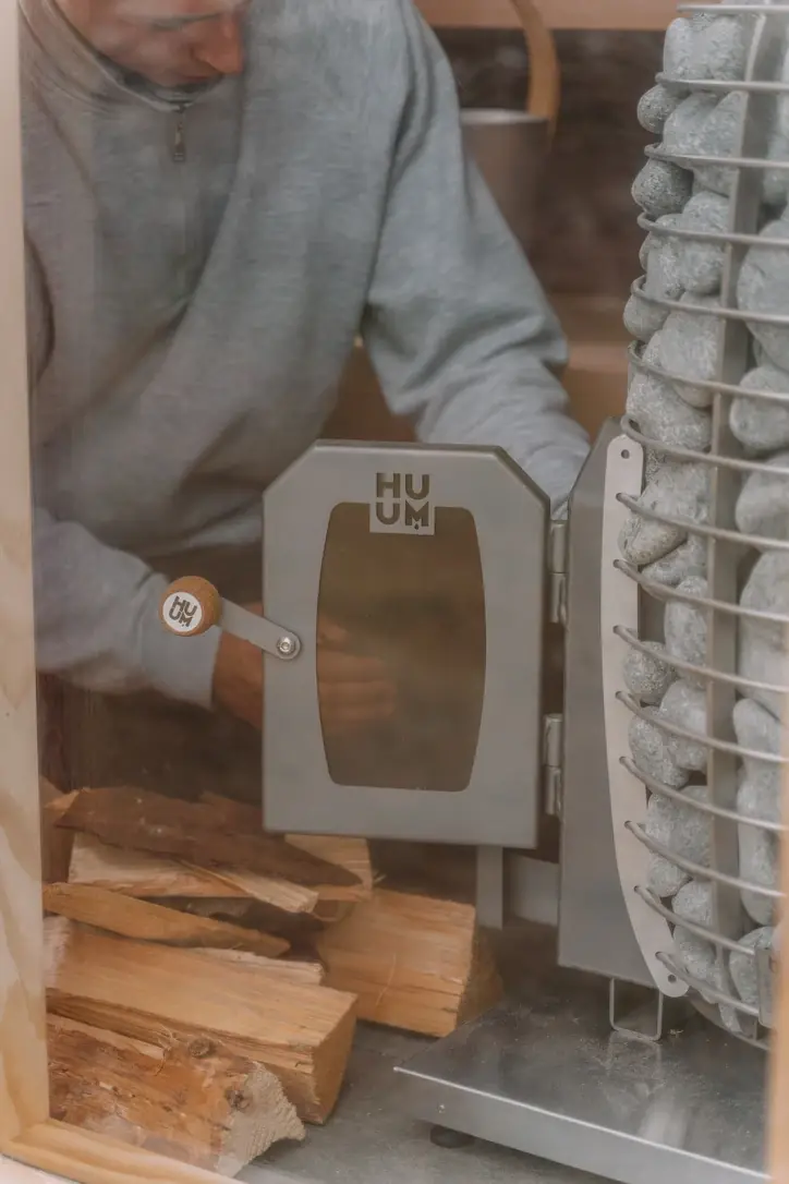 A person tending a wood-fired sauna heater surrounded by stones